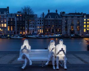 Installazione di light art con tre giovani che guardano fissi lo schermo di un telefonino seduti su una panchina